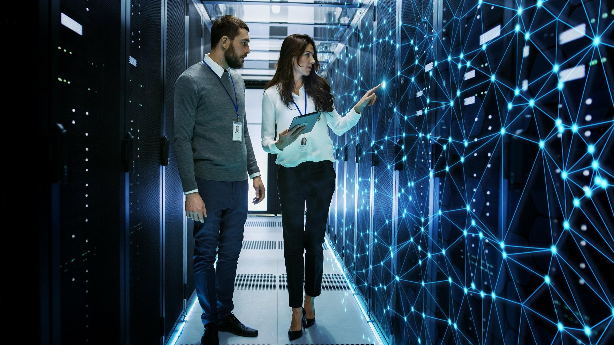 two people in an aisle between computing racks, pointing at interconnected dots overlaid on a supercomputer